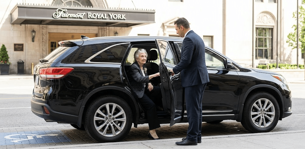 Professional chauffeur assisting an elderly woman into a luxury black SUV in front of the Fairmont Royal York Hotel in Toronto.